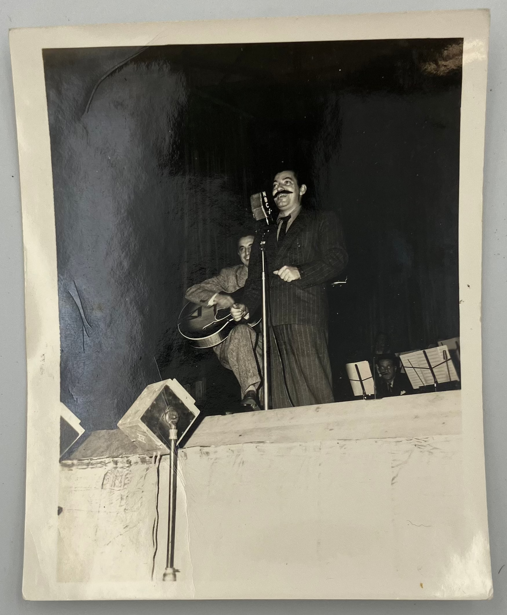 A vintage black and white photograph print depicting a performer on stage with a microphone and guitar, displayed against a neutral gray background.