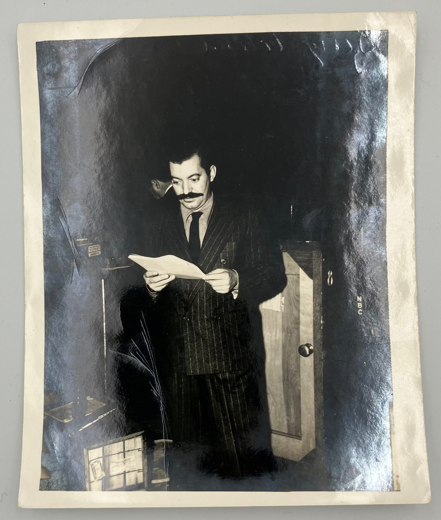 A vintage black and white portrait photograph print of a man in a pinstripe suit holding a piece of paper, displayed on a neutral gray background.