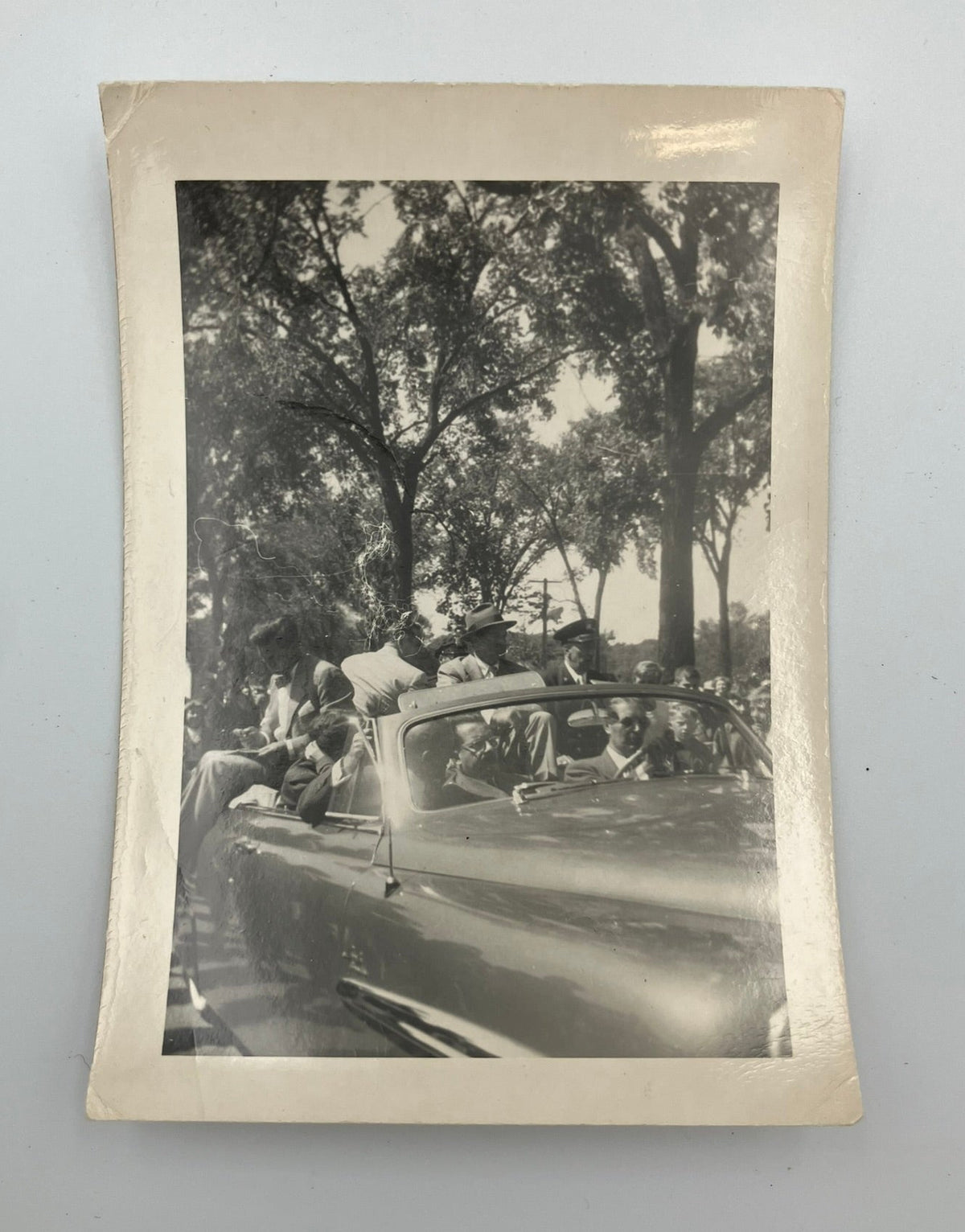 A vintage black and white photograph print with a cream-colored border, depicting a group of people riding in a convertible car under trees, displayed against a light gray background.