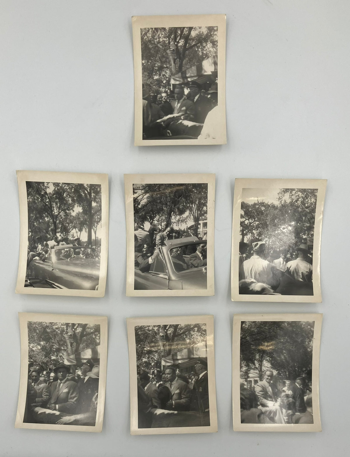 Seven vintage black and white photographs with aged cream borders arranged on a light gray wall, depicting people in old cars and outdoor scenes.