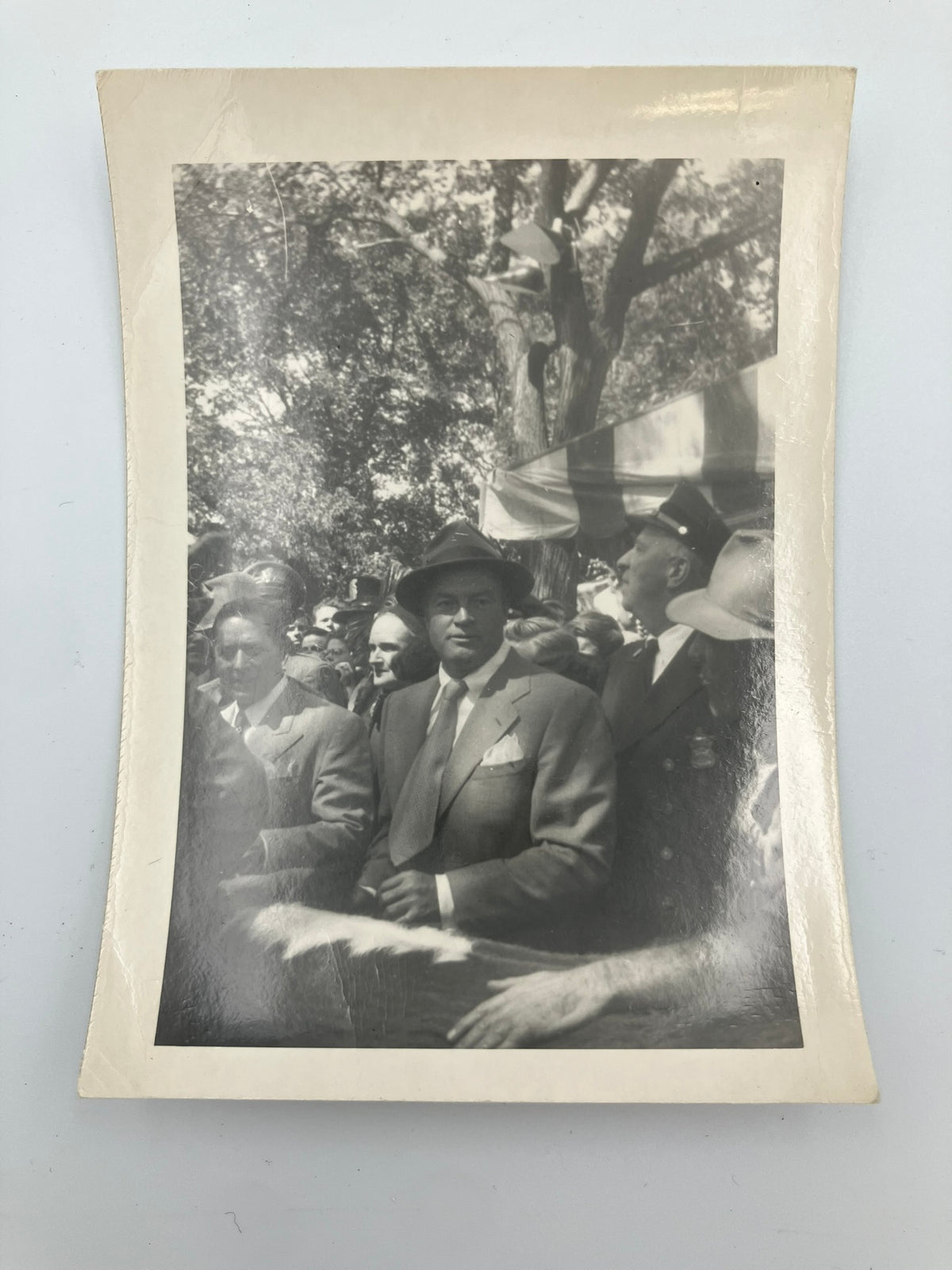 A vintage black and white photograph print with a cream-colored border, depicting a historical crowd scene with a man in a suit and hat.