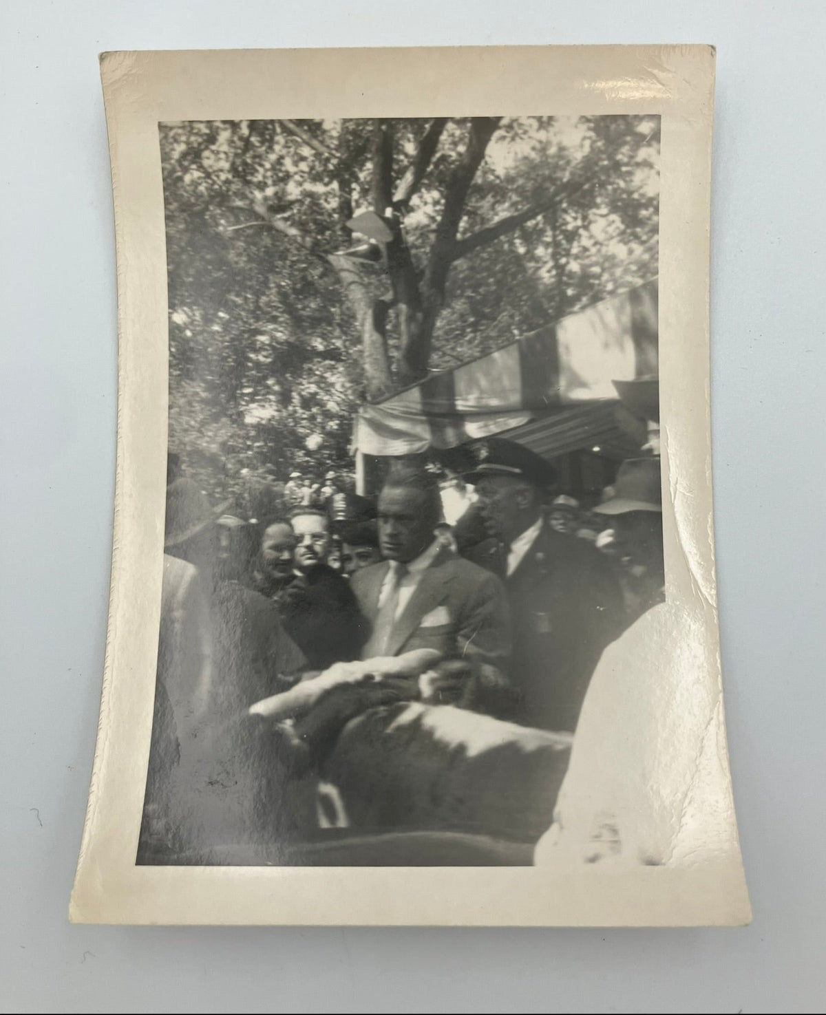 A vintage black and white photograph print with a cream-colored border, showing a historical outdoor scene with trees and people, displayed against a light gray background.