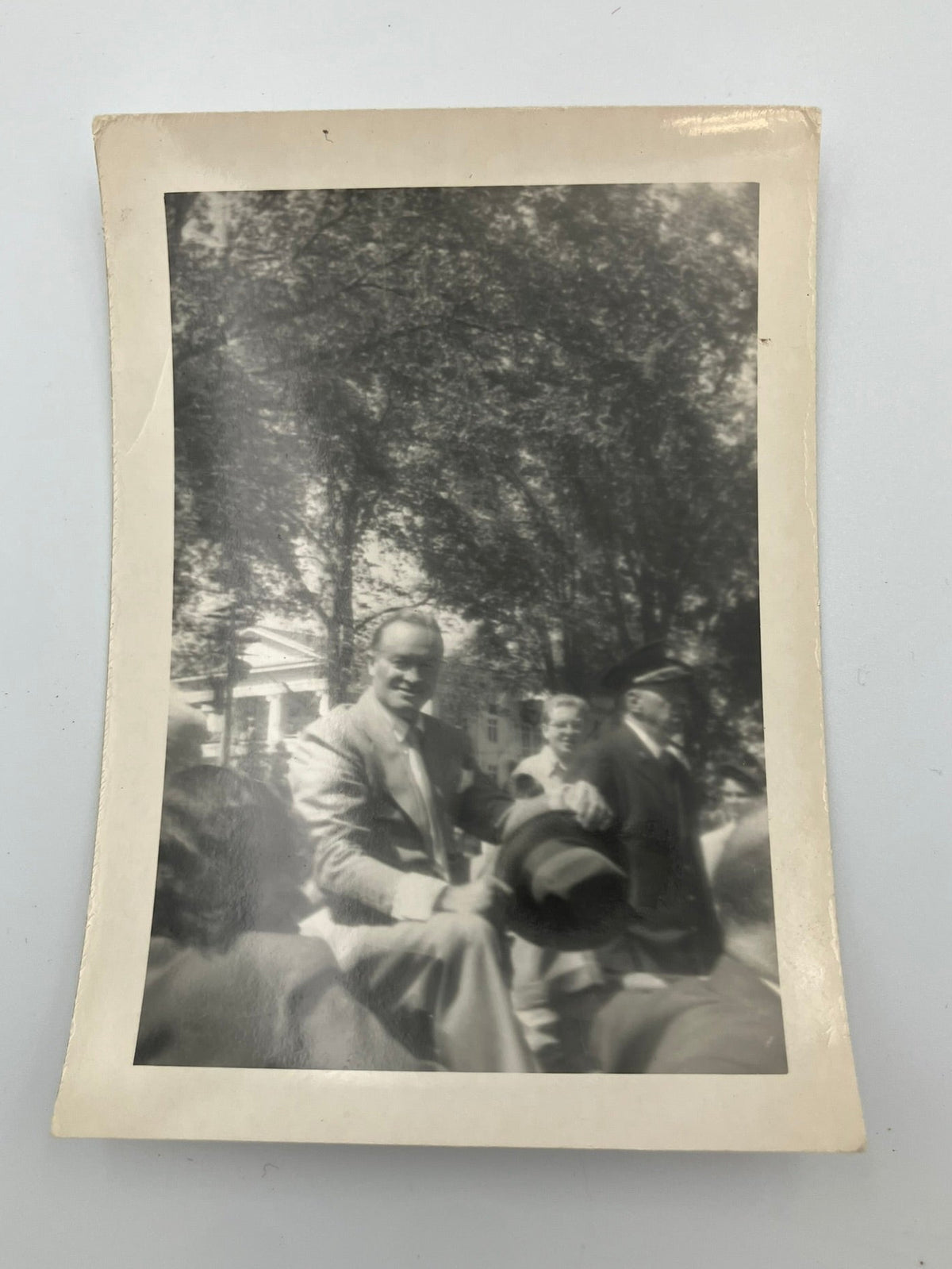 A vintage black and white photograph print with a cream-colored border, depicting a group of men outdoors under trees.
