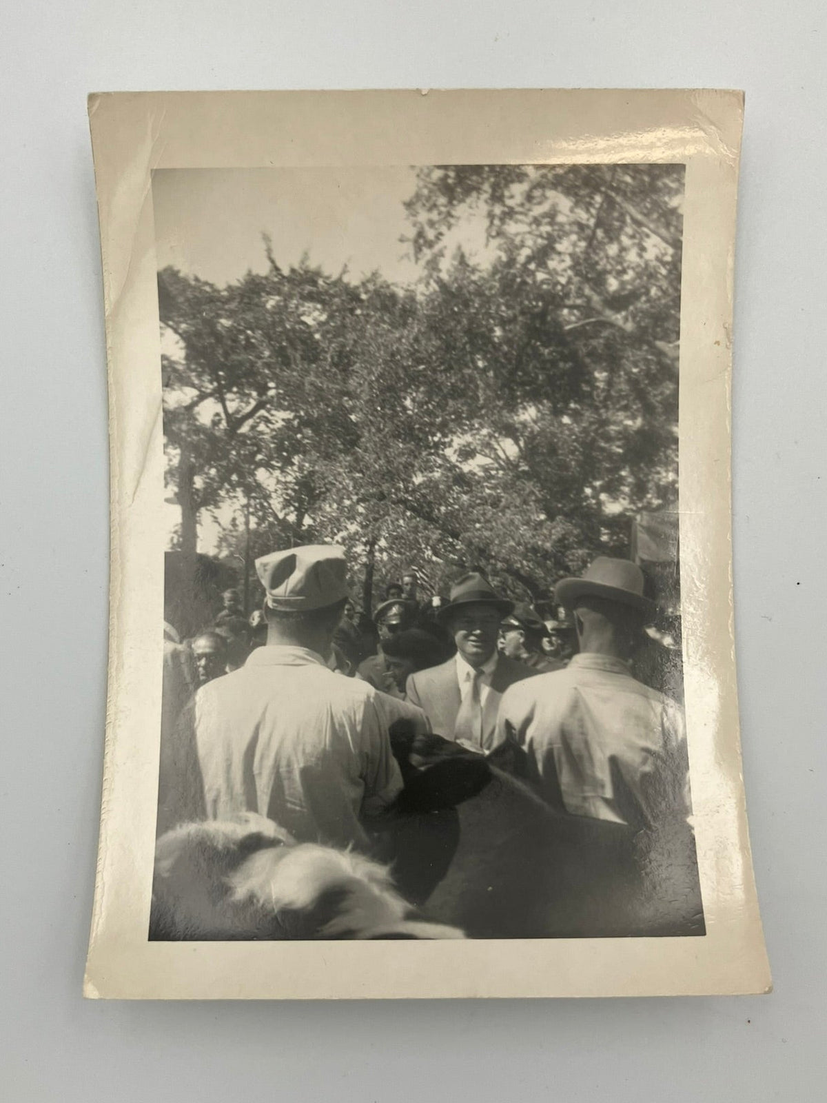 A vintage black and white photograph print with a cream-colored border, depicting a group of men outdoors under trees, displayed against a light gray background.