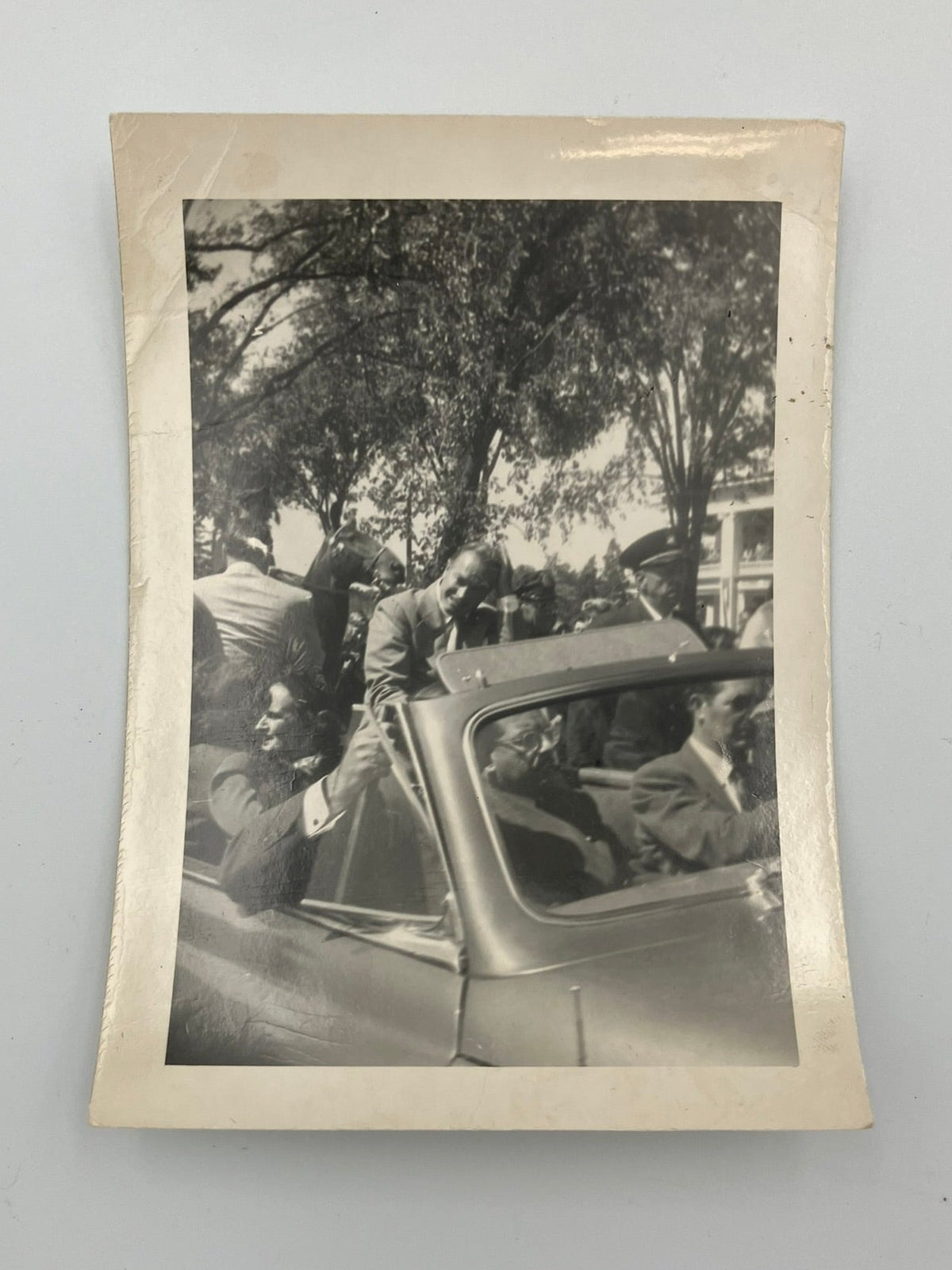 A vintage black and white photograph print with a cream-colored border, depicting a crowd scene with people in and around an old car, displayed against a light gray background.