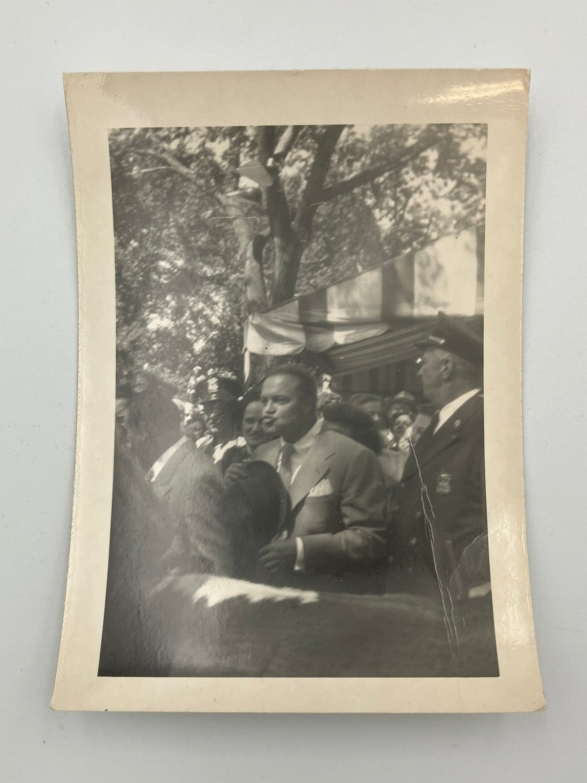 A vintage black and white photograph print with a beige border, depicting a historical outdoor scene with a man in a suit standing in front of trees and a building.