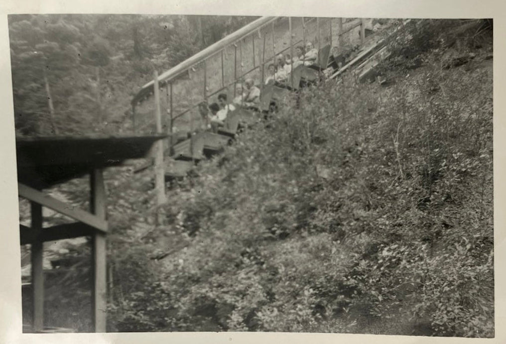 A vintage black and white photograph print with a scalloped border, depicting an outdoor scene with trees and a wooden structure, shown against a neutral gray background.