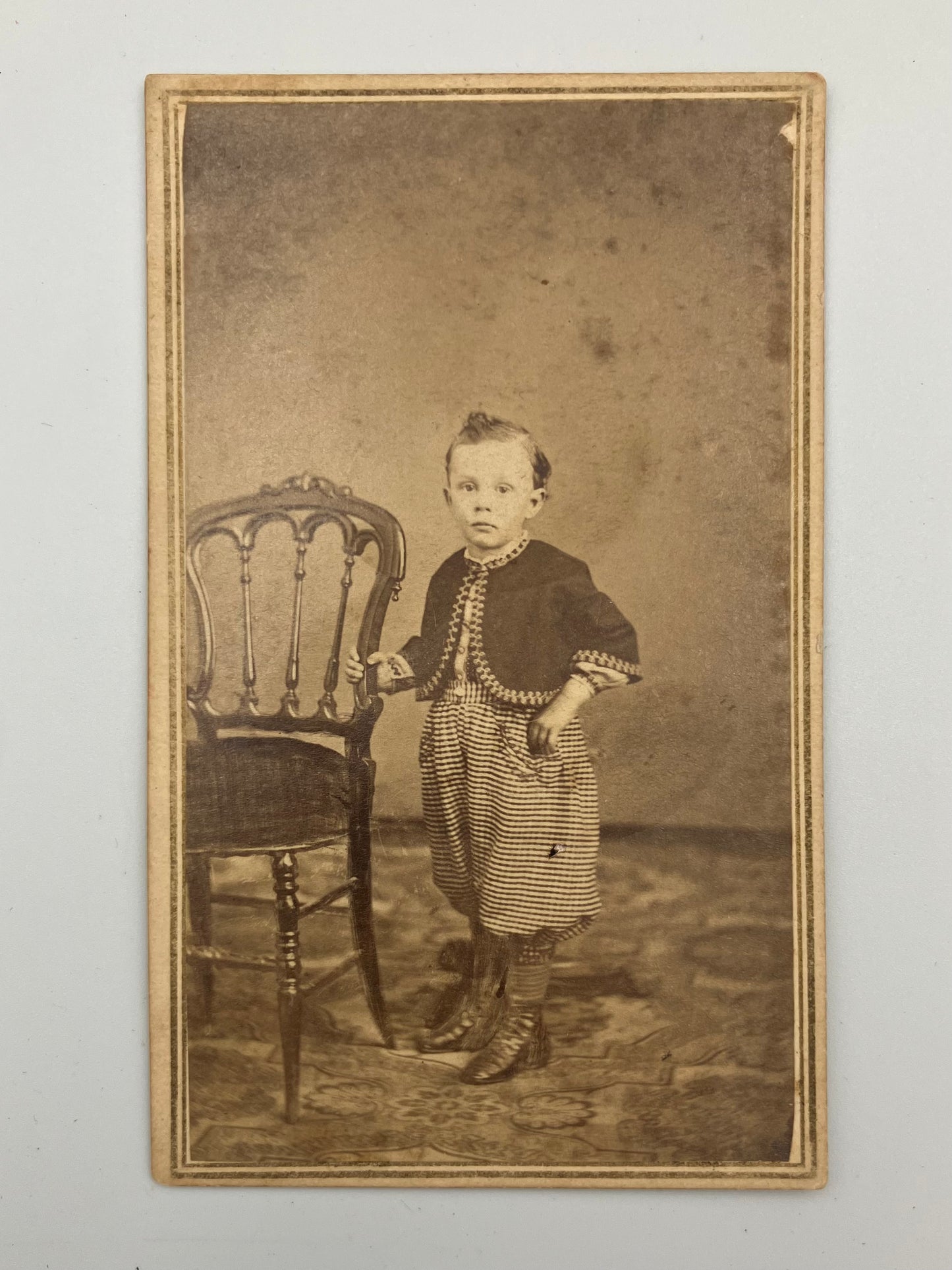 An antique sepia-toned carte de visite photograph showing a young child standing beside a chair against a mottled studio backdrop, with a decorative border around the print.