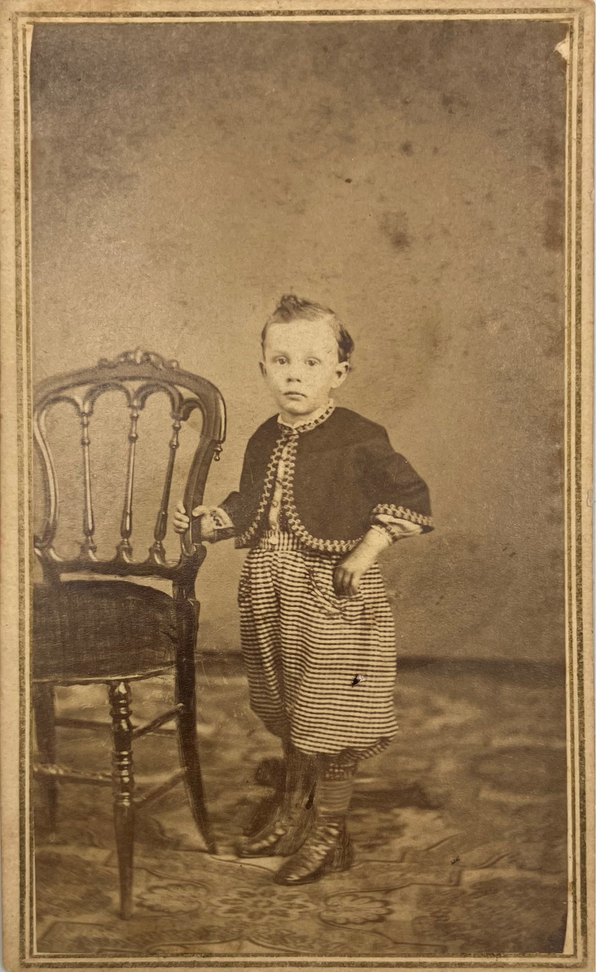 Vintage sepia-toned portrait photograph of a young child standing beside a wooden chair against a mottled studio backdrop.