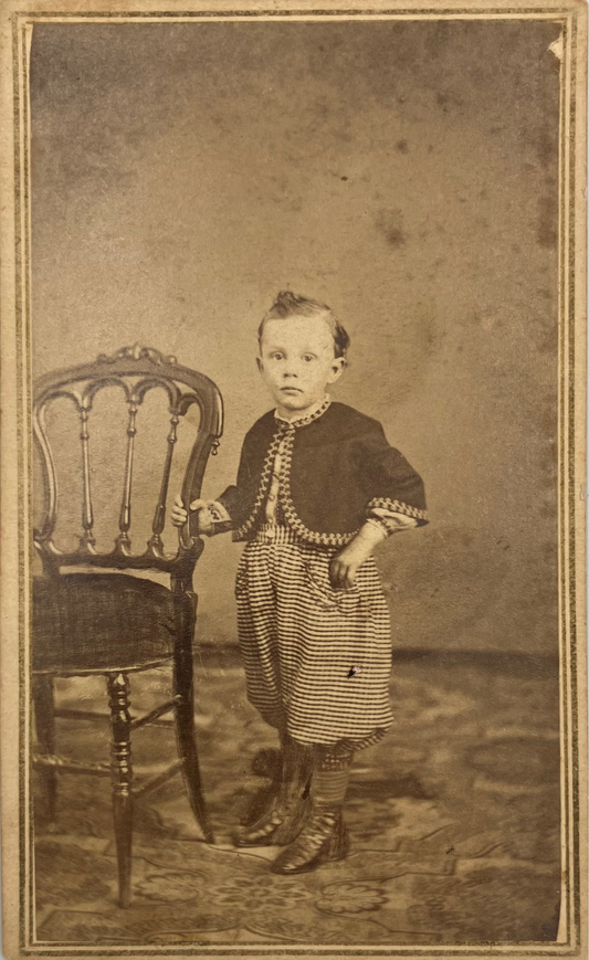 Vintage sepia-toned portrait photograph of a young child standing beside a wooden chair against a mottled studio backdrop.