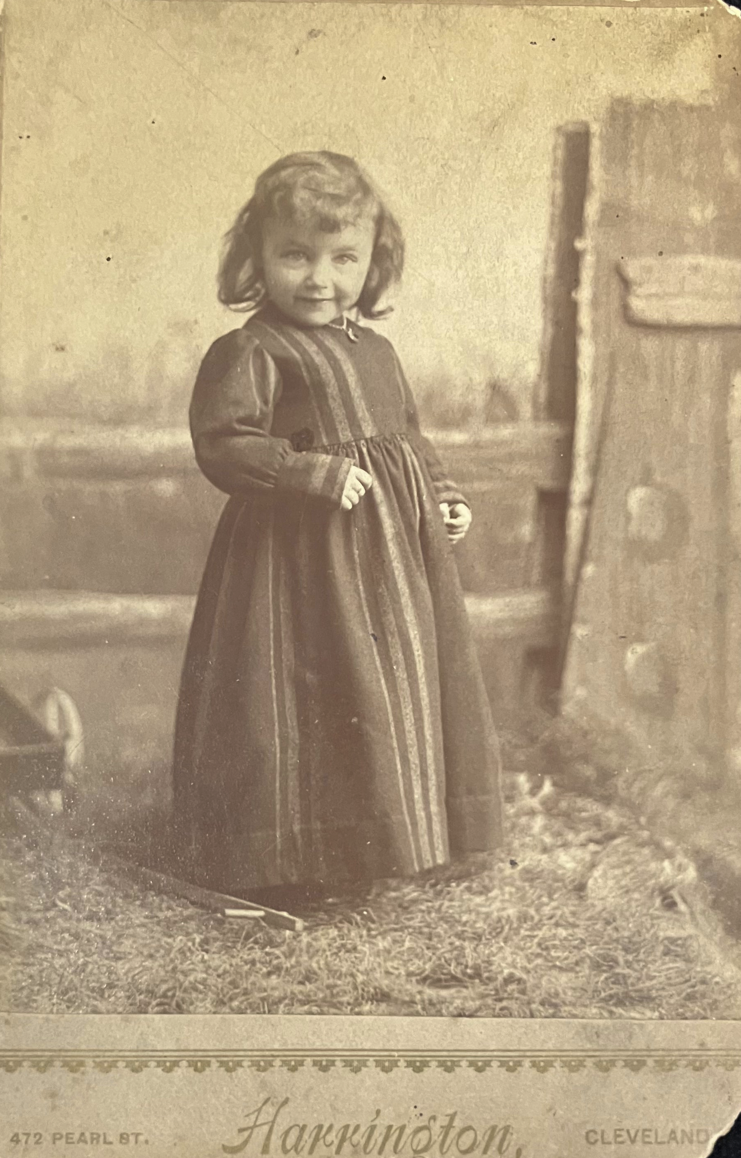 Vintage sepia-toned photograph of a young girl in a dress standing outdoors.