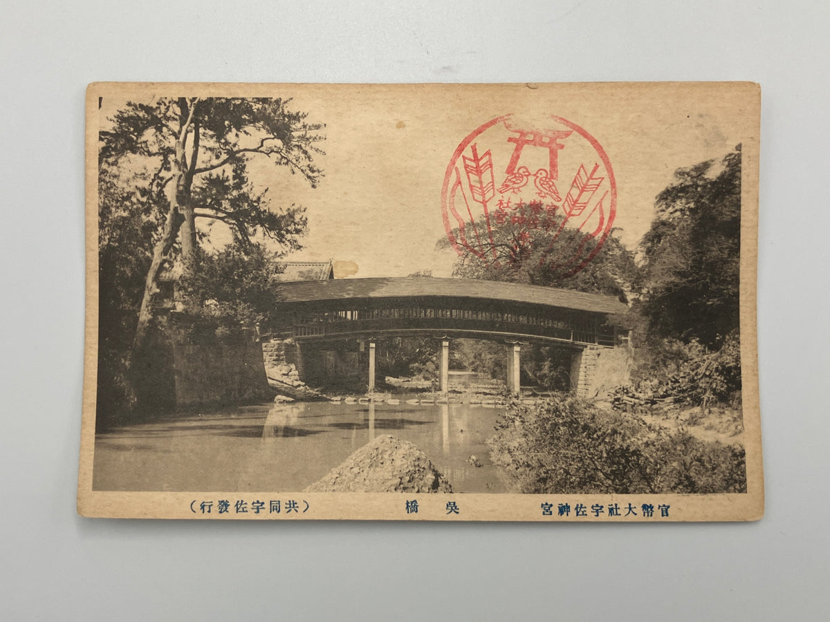 A vintage Japanese postcard showing a covered bridge over water, with trees on both sides and a red circular stamp across the upper right area. The postcard has Japanese text printed along the bottom and is photographed against a light gray background.