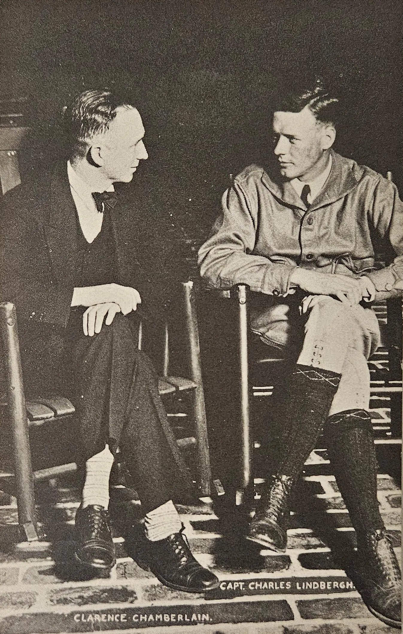 Two men sitting on chairs, one in formal attire and the other in military uniform, with a brick floor and wooden chairs in the background.