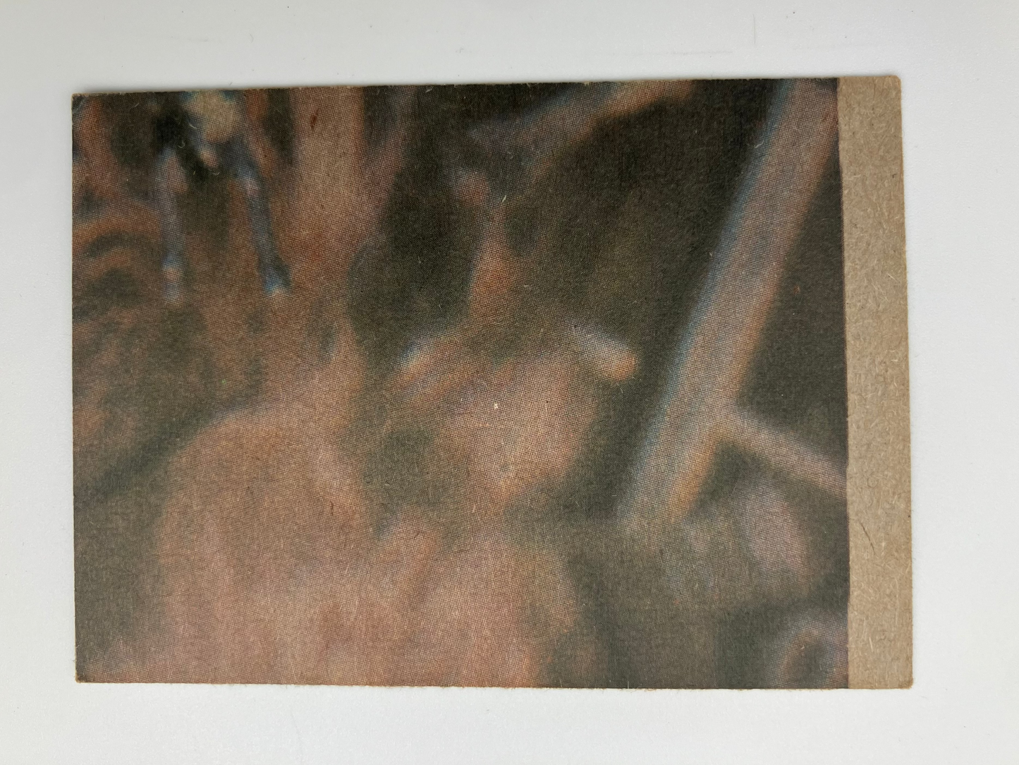 A square art print or photograph mounted on a light gray background, featuring an abstract close-up scene in earthy tones of brown, tan, and dark gray. The print has a textured surface and a tan strip along the top edge.
