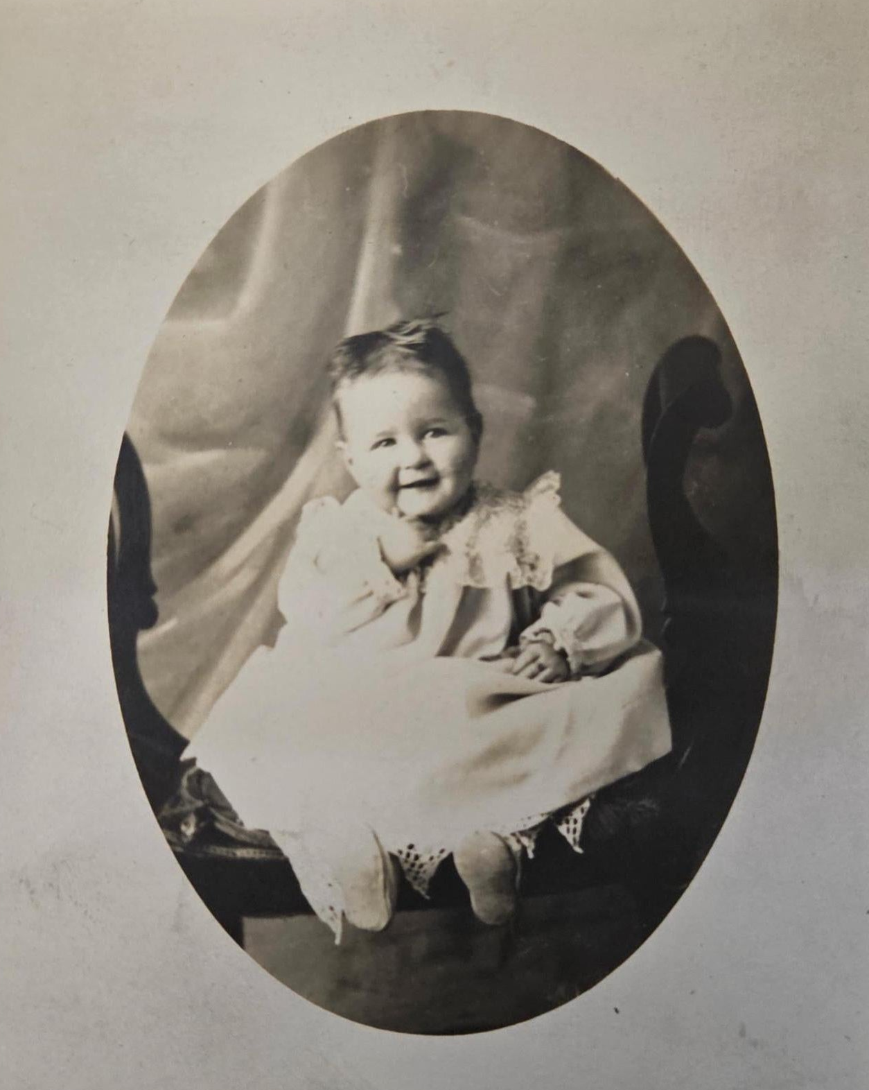 Oval shaped black and white portrait photo of a young child, a smiling baby Evelyn, seated on a bench.