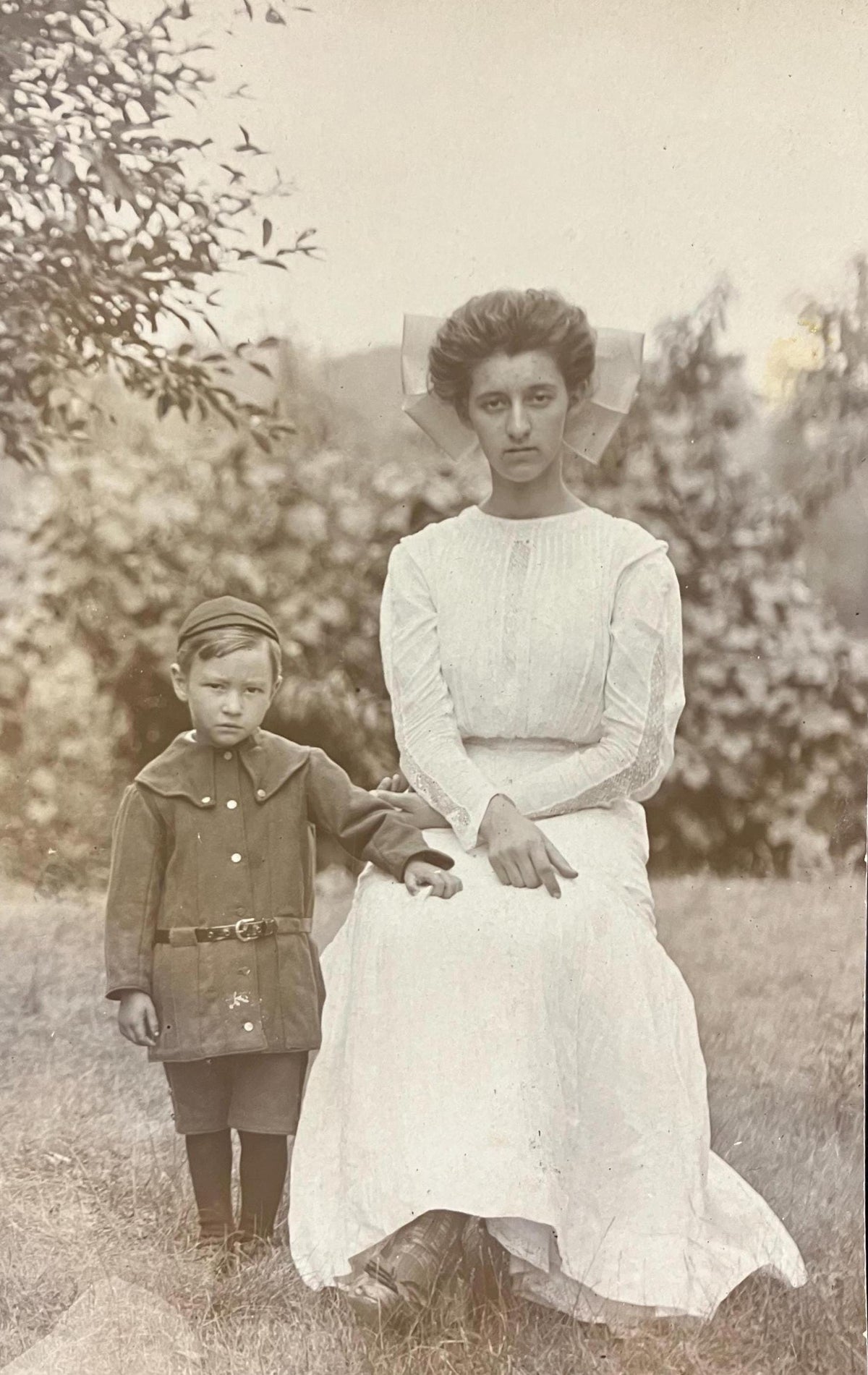 A vintage black and white photo of a mother and son dressed up in formal white and suit, respectively, with somber expressions, likely from the early 20th century.