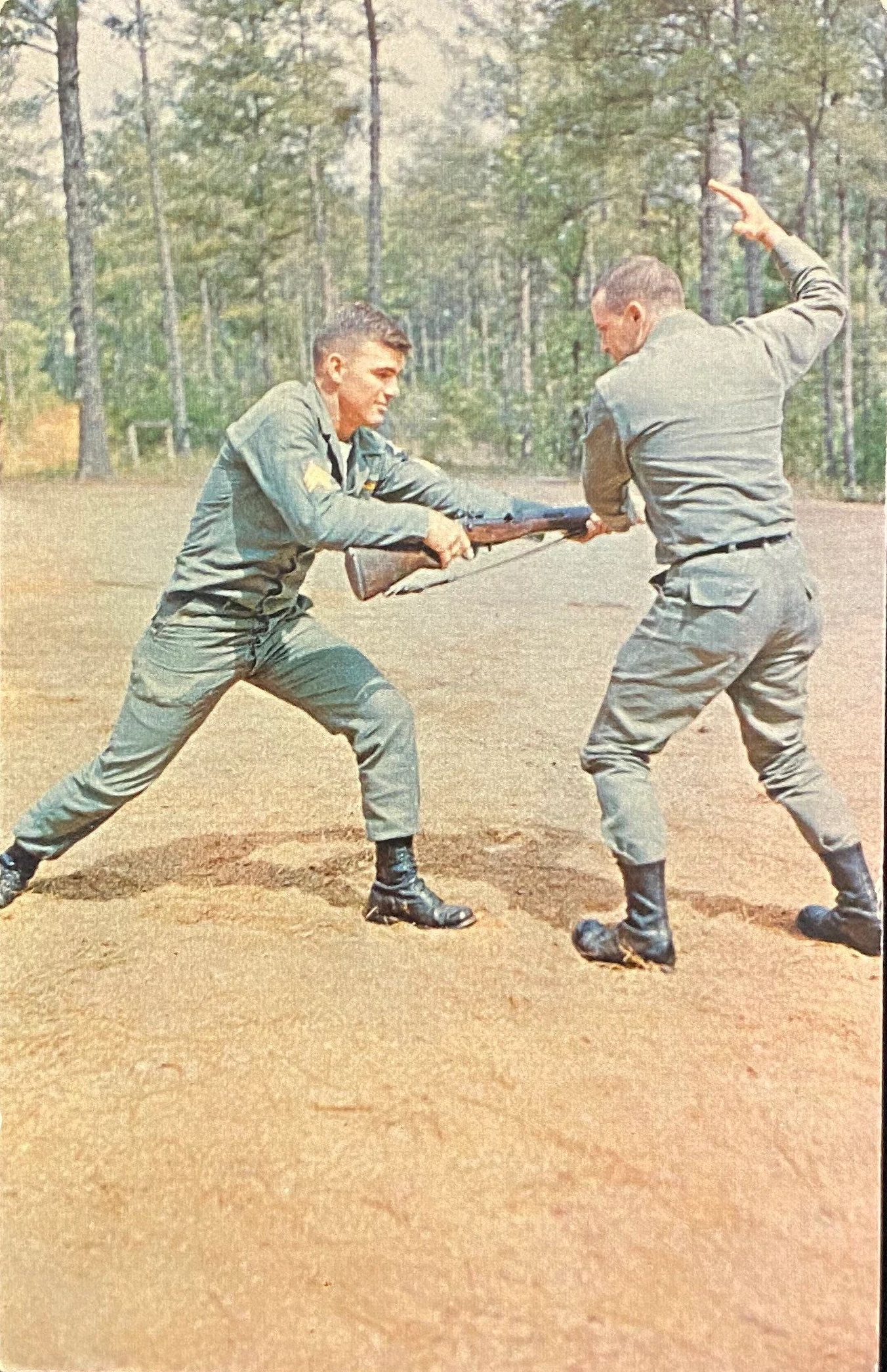 Two soldiers in uniforms practicing hand-to-hand fighting with a rifle on a dirt field.