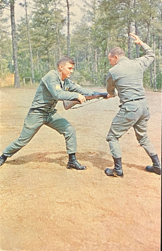 Two soldiers in uniforms practicing hand-to-hand fighting with a rifle on a dirt field.