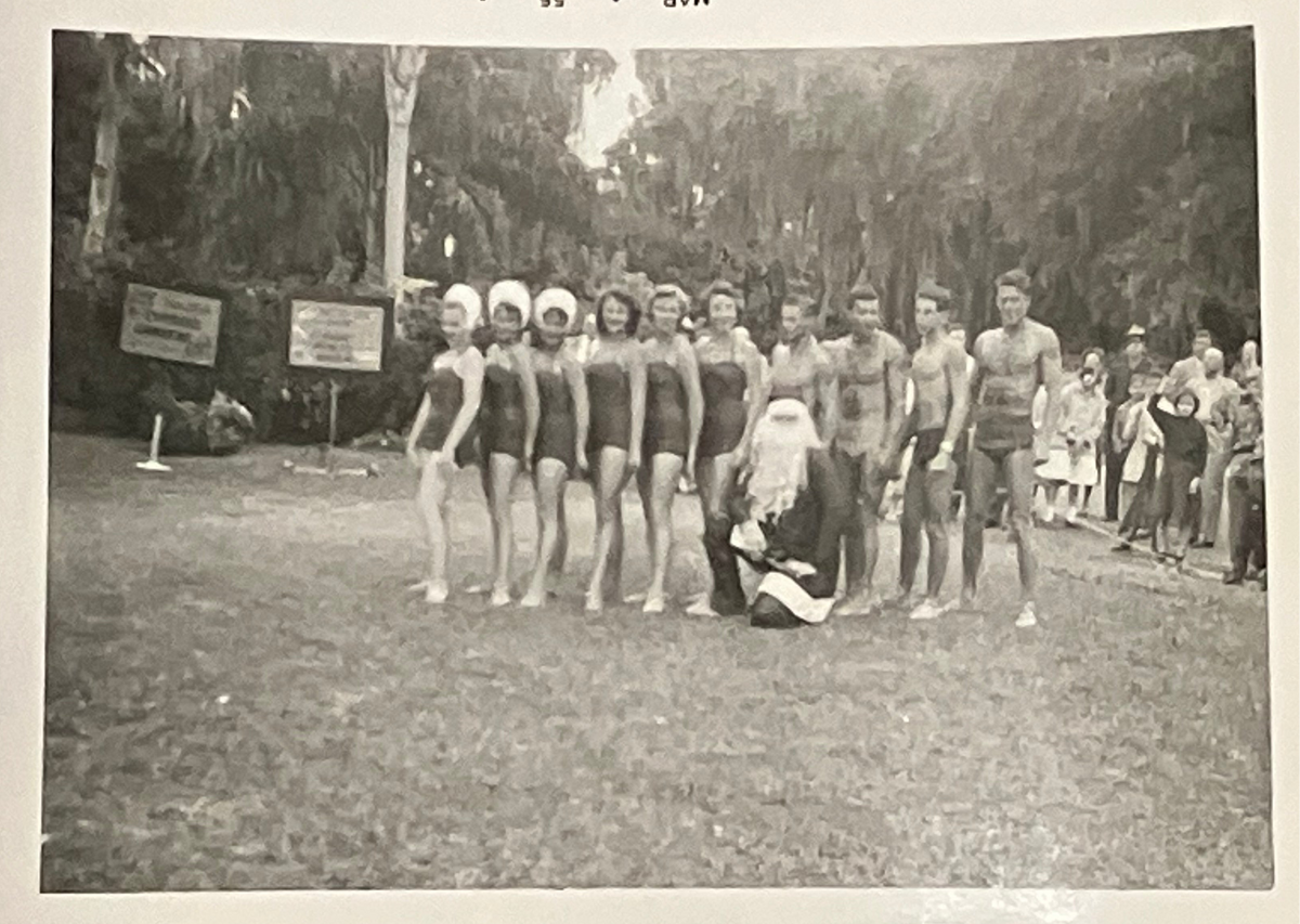 Vintage black and white photo of a line of people in bathing suits standing on a grassy field. Date on the frame is Mar 55.